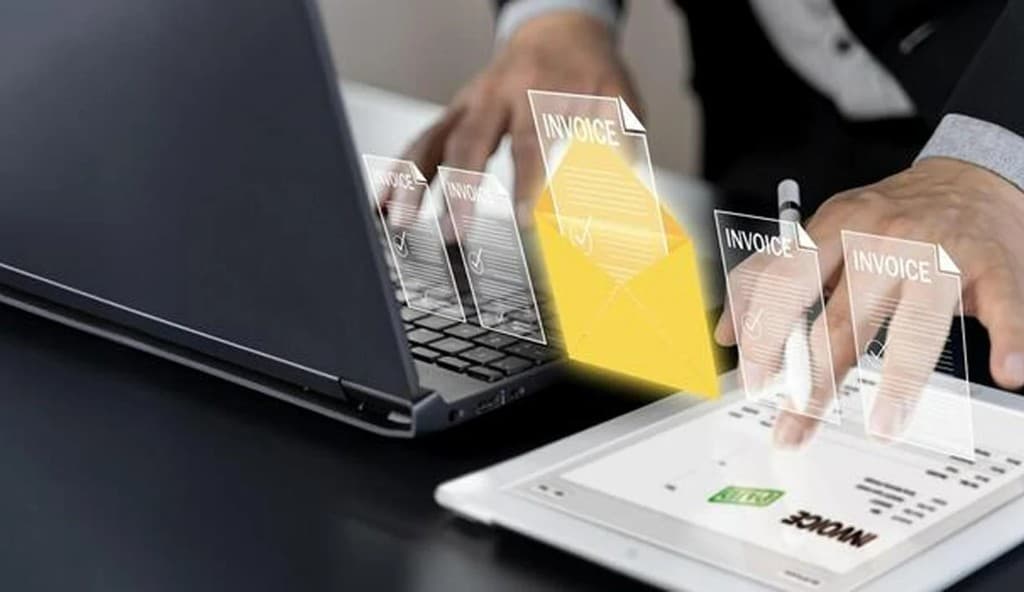 A person using a laptop and tablet with a glowing invoice envelope overlay